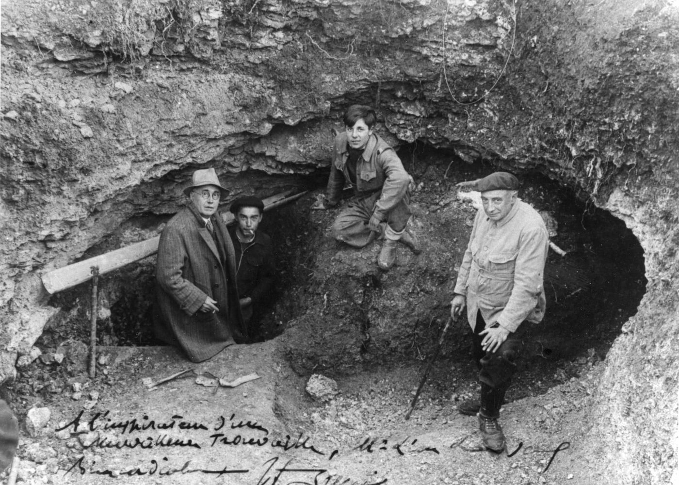 L’entrée de la grotte de Lascaux en septembre 1940 : trois jeunes gens et un homme en manteau posent autour du trou ouvert dans la pente, sur un tirage dédicacé
