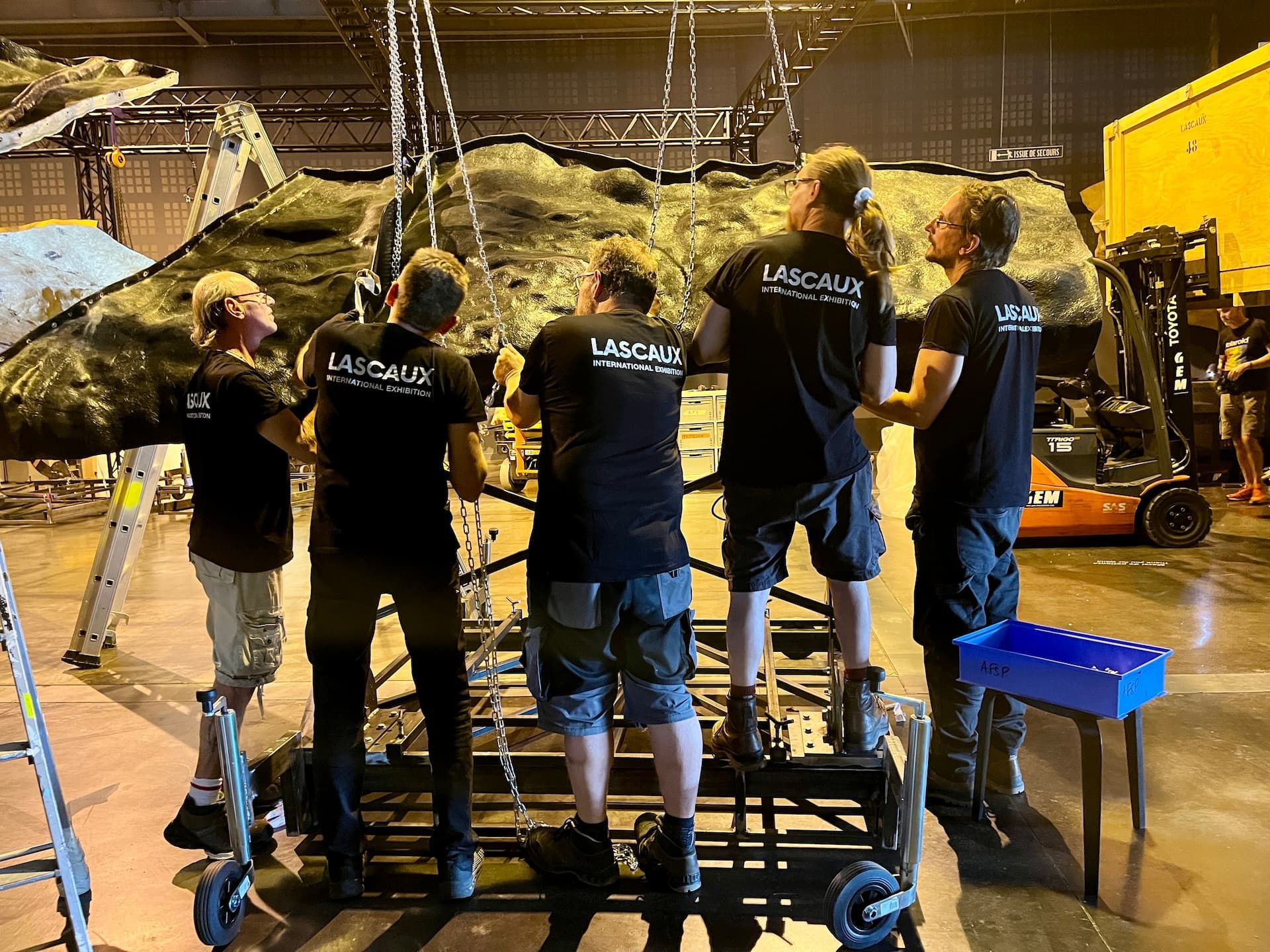 Five team members around a section of painted wall hanging from its hoist, in the assembly hall